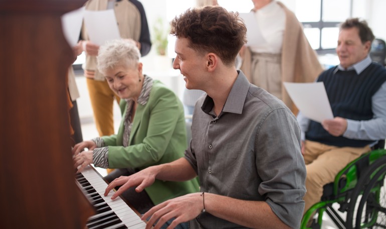 Ung man spelar piano tillsammans med en äldre kvinna på ett äldreboende.