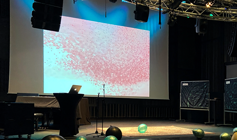 En konsertsal med en projektorduk i bakgrunden. Ballonger ligger på golvet.