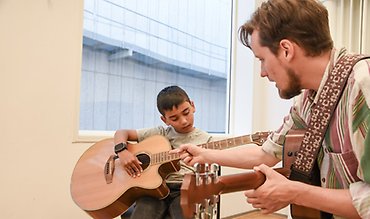 En övningselev och en lärarstudent sitter i ett rum snett vända mot varandra. Övningseleven spelar på en stor gitarr medan han får instruktioner av lärarstudenten. 
